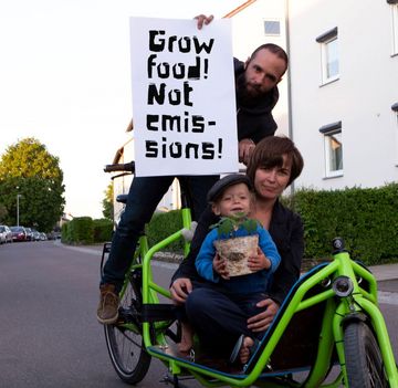 Eine Familie sitzt mit einer TOmatenpflanze auf einem Lastenrad und hält ein Demo-Schild mit dem englischen Spruch: Grow food - not emissions