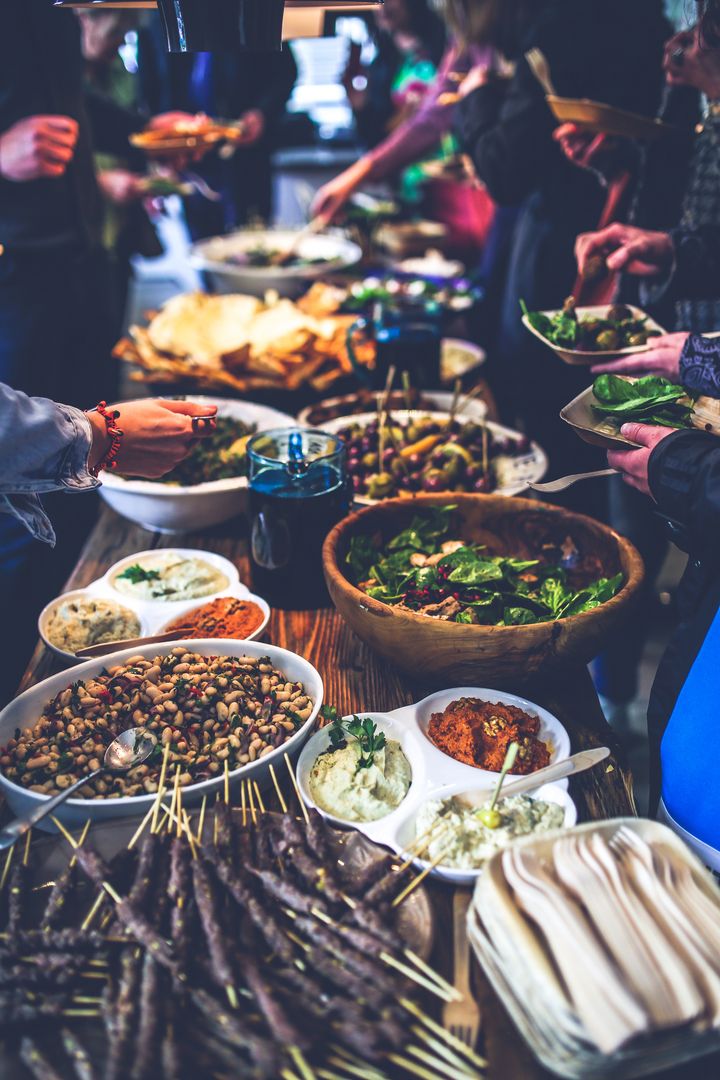 Auf einem großen Tisch steht eine Vielfalt an lecker aussehen Speisen, drum herum viele gesellige Menschen